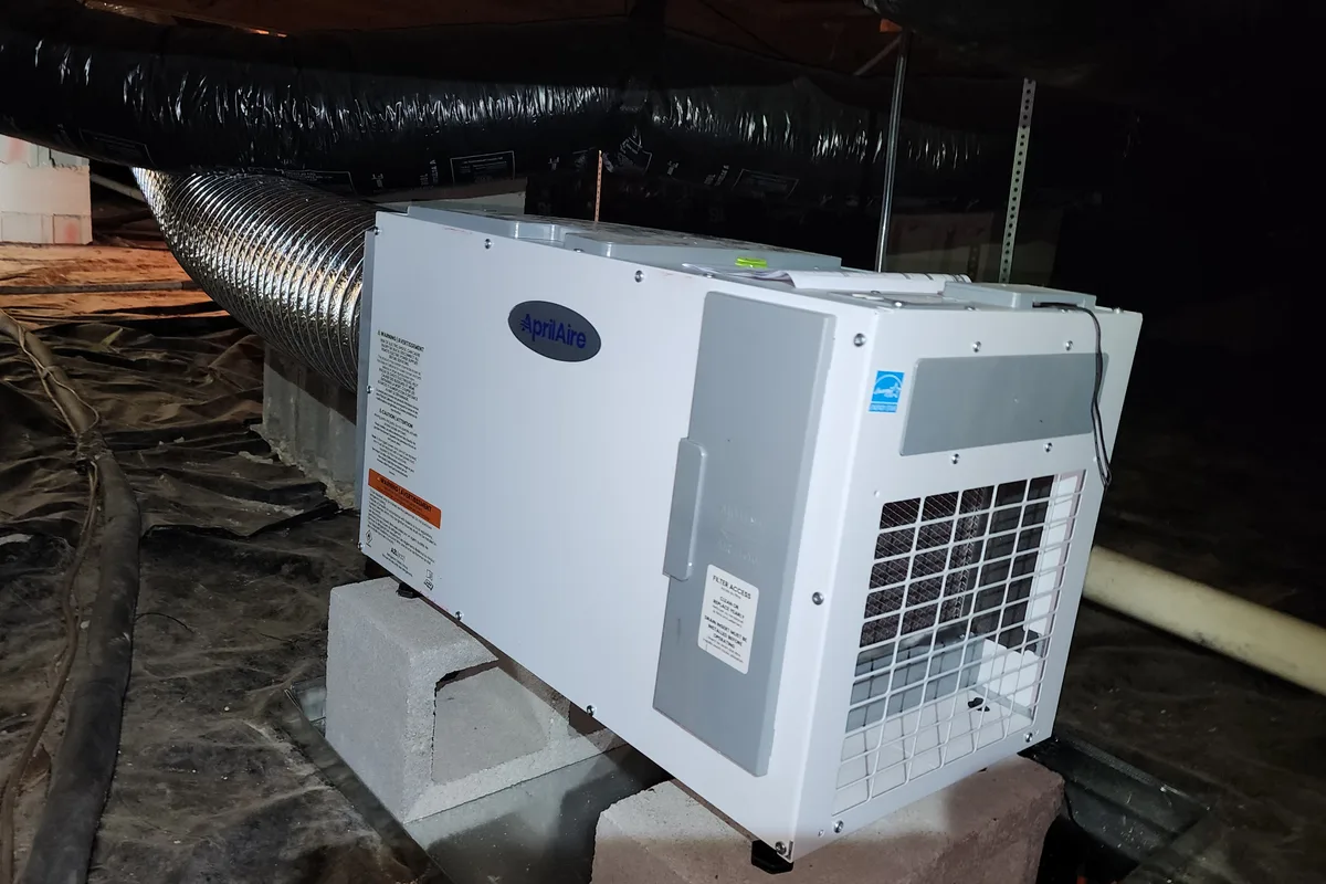 AprilAire dehumidifier mounted on concrete blocks in a crawl space with black vapor barrier, flex ductwork, and PVC piping visible, Wake County NC