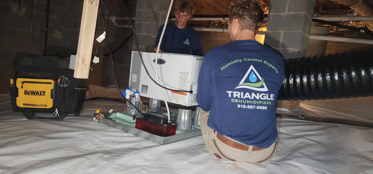 Two Triangle Dehumidifiers technicians installing an AprilAire dehumidifier on support blocks in an encapsulated Wake County NC crawl space, condensate pump and ductwork visible