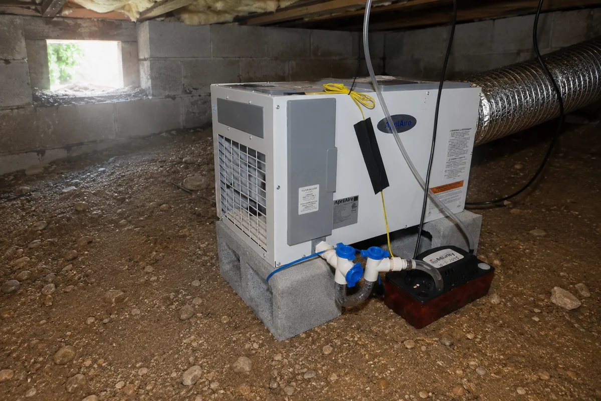 AprilAire crawl space dehumidifier installed in a vented crawl space beneath a Raleigh NC home, condensate line running toward the foundation perimeter
