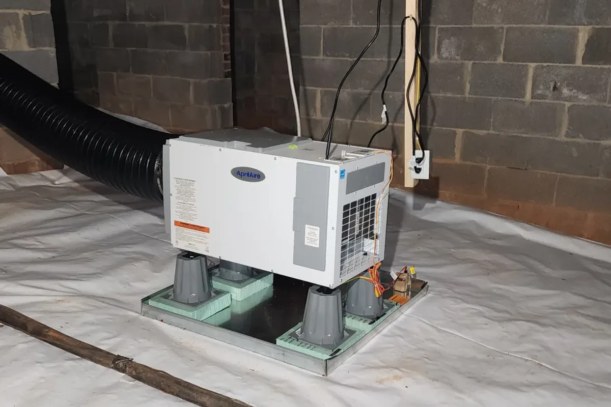 AprilAire crawl space dehumidifier installed on a drain pan over a white ground vapor barrier in a vented crawl space with block foundation walls in Wake County NC