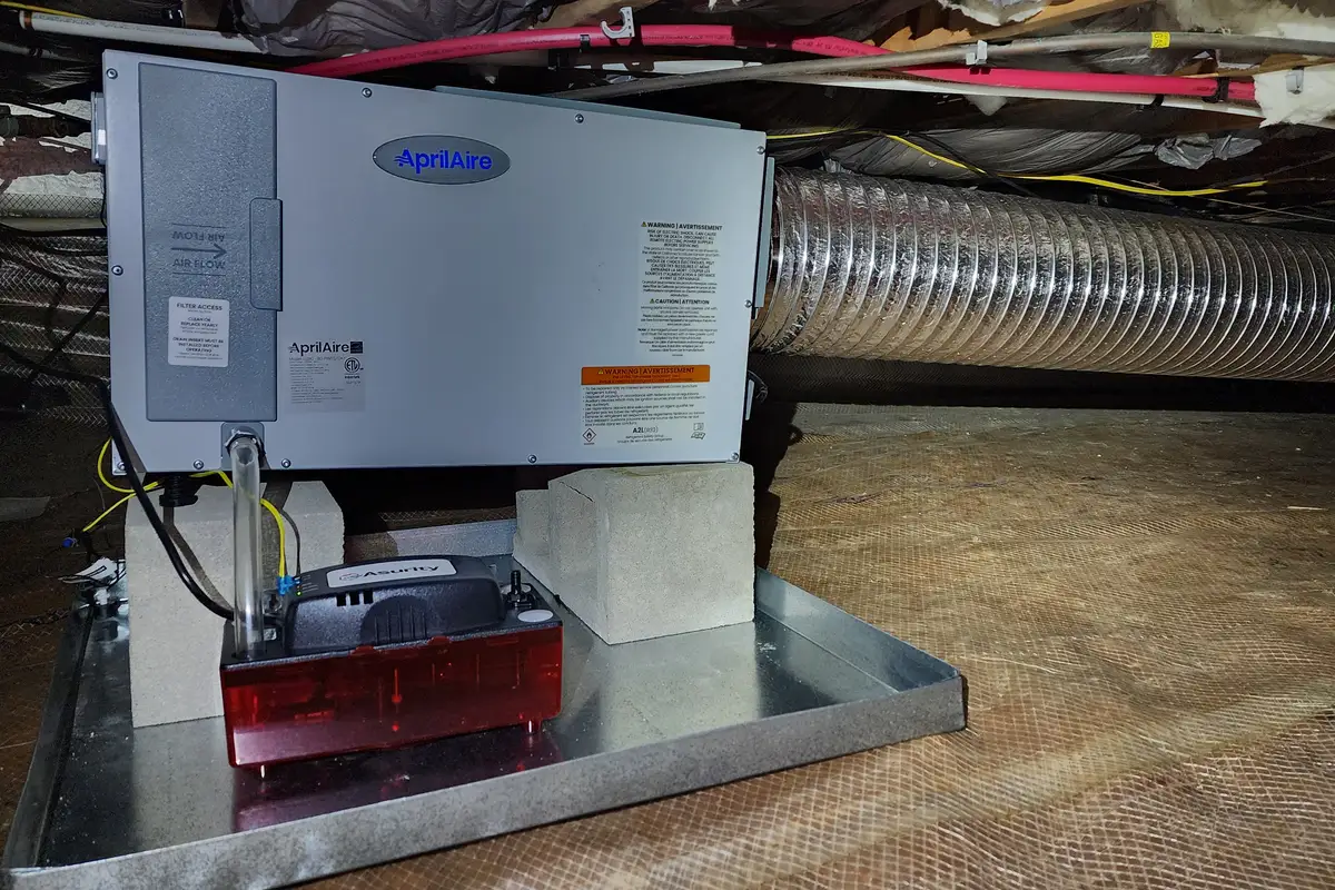 AprilAire crawl space dehumidifier installed on a metal drip pan in an Apex NC crawl space, with condensate pump and flexible duct visible above a vapor barrier