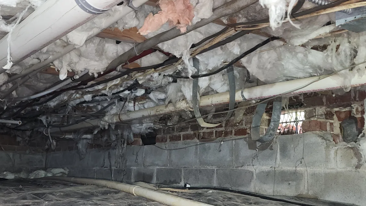 Problem crawl space in the Triangle NC showing damaged fiberglass insulation falling from floor joists, exposed pipes and wiring, and an open foundation vent in a brick and block wall
